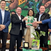 Um grupo de cinco pessoas está posando para uma foto em frente a um banner colorido com ilustrações de trabalhadores rurais. No centro, uma mulher está segurando um certificado ou documento junto com os dois homens ao seu lado. Todos estão sorrindo. A mulher está usando um blazer branco e calças bege, e os homens estão vestindo trajes formais, como ternos. Em uma mesa à frente deles, há uma planta e alguns itens decorativos.