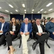 Um grupo de pessoas está sentado em uma fileira de cadeiras em um auditório ou sala de conferências. A foto foi tirada de frente para eles. As pessoas estão vestidas de maneira formal e parecem estar participando de um evento. Há várias outras pessoas sentadas e conversando ao fundo. Um banner ao fundo menciona "Solidariedade ao Povo Gaúcho" e outras frases relacionadas ao evento.